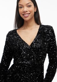 Jauna sieviete smaidot, ģērbusies garām piedurknēm melnā kleitā, kas ir pārklāta ar spīdīgiem melniem miroņiem, uz vienkārša balta fona.