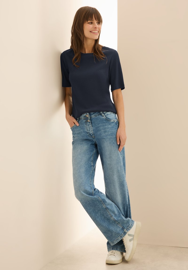 Haut à manches courtes bleu marine, coupe décontractée. Jean large déchiré bleu clair. Baskets blanches avec des accents gris. Fond neutre.