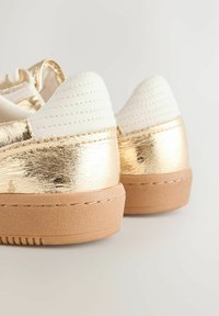 Gold metallic sneakers with white textured accents, featuring a smooth finish and a chunky tan sole, showcasing detailed stitching on the heel.