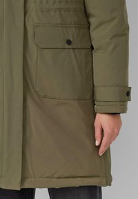 Olijfgroene parka met een gestructureerd oppervlak, voorzien van een knoopsluiting flapzak en manchetten, vergezeld van een glad, gestroomlijnd ontwerp.