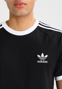 Muž v černém tričku Adidas s bílým límečkem, bílými manžetami a třemi bílými pruhy na ramenou.
