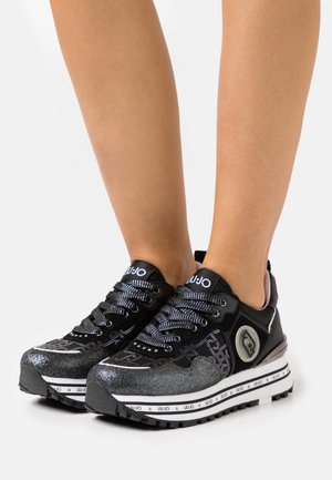 Baskets basses noires et argentées avec des lacets à motifs et des semelles épaisses blanches de marque, portées sur des jambes nues.