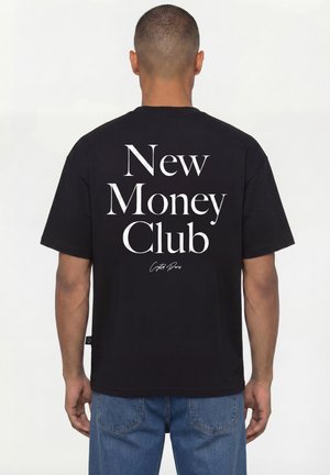 Mann trägt ein schwarzes T-Shirt mit weißem Schriftzug „New Money Club“ auf dem Rücken, kombiniert mit blauer Jeans, steht vor weißem Hintergrund.