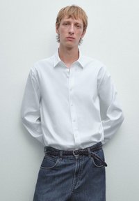 Weißes Hemd mit langen Ärmeln, kombiniert mit dunklen Jeans und einem geflochtenen schwarzen Gürtel. Das Model steht vor einem hellen Hintergrund.
