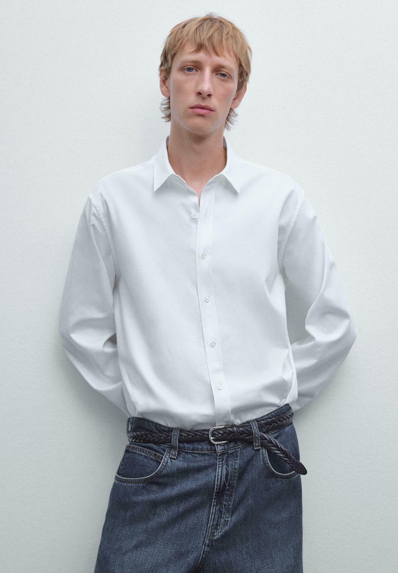 Weißes Hemd mit langen Ärmeln, kombiniert mit dunklen Jeans und einem geflochtenen schwarzen Gürtel. Das Model steht vor einem hellen Hintergrund.