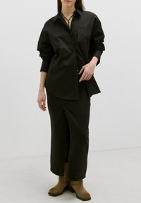 Schwarzhemd mit einer Tasche, kombiniert mit einem schwarzen Jeansrock mit einem seitlichen Schlitz. Braune Knöchelstiefel mit einem dicken Absatz runden das Outfit ab.