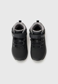 Un par de zapatillas negras para niños con forro gris interior, cordones elásticos y tiras de Velcro sobre un fondo blanco.