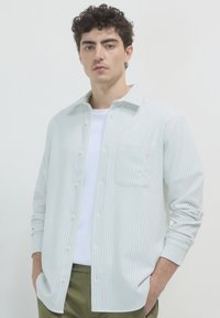 Un jeune homme avec des cheveux bouclés et foncés porte une chemise blanche à rayures sur un T-shirt blanc, avec les mains dans les poches d'un pantalon vert olive.