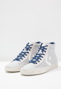 Vysoké tenisky vyrobené z světle šedé tkaniny s bílým logem hvězdy, modrými tkaničkami a bílými gumovými podrážkami. Texturovaný plátěný svršek.