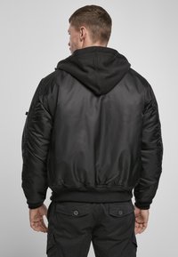 Giacca bomber nera con finitura liscia, polsini e orlo a coste, con cappuccio. Maniche strutturate con dettagli arricciati.