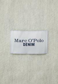 Uno sfondo di tessuto bianco con un motivo testurizzato. Un'etichetta rettangolare blu e grigia riporta la scritta "Marc O'Polo DENIM" in lettere blu scuro.