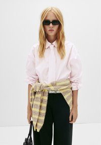 Cămașă în dungi roz și alb, cardigan legat în dungi galbene și gri, pantaloni negri evazați și ochelari de soare mici negri. Design minimalistic.