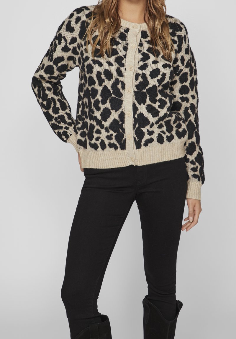 Femme portant un cardigan boutonné imprimé léopard beige et noir, un pantalon noir et des bottes noires, debout avec la main sur la hanche.