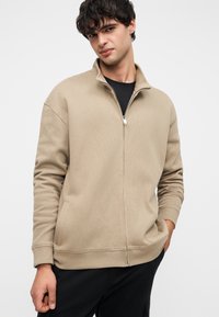 Μπεζ φερμουάρ φούτερ με ψηλό γιακά, υφή ύφασμα, πλευρικές τσέπες και ριμπ μανσέτες. Φορεμένο πάνω από μαύρο πουκάμισο.