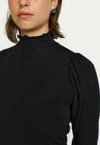 Črn zgornji del z visokim ovratnikom iz teksturiranega materiala, s temi napihnjenimi rokavi in nabranim ovratnikom. Gladka tekstura in prilegajoča zasnova.