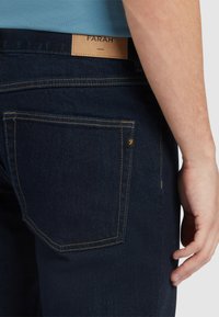 Jeans in denim scuro con un design a gamba dritta, cuciture gialle e una patch in pelle con l'etichetta "FARAH" in vita, che mostra una tasca posteriore.