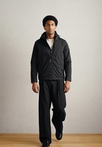 Μαύρο μπουφάν με διακοσμητική ραφή και φερμουάρ μπροστά, σε συνδυασμό με μαύρο παντελόνι φαρδιάς γραμμής. Μινιμαλιστικός σχεδιασμός, φορεμένο πάνω από λευκή ριπ μπλούζα.