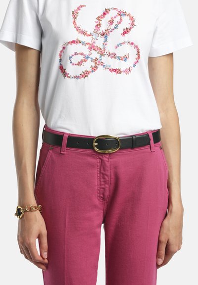 Wit t-shirt met kleurrijk bloemenpatroon, ingestopt in een hooggesneden magenta broek, zwarte riem met gouden gesp en een gouden armband om de linkerpols.
