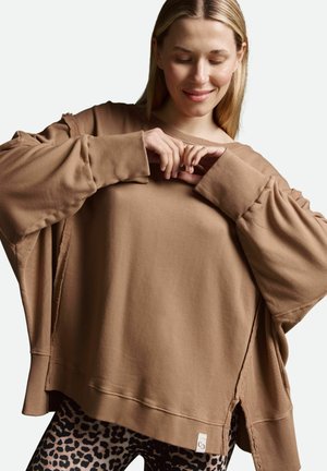 Brauner oversized Pullover mit abfallenden Schultern, gerippten Bündchen und getragenen Akzenten, kombiniert mit schwarzen Leggings im Leopardenmuster.