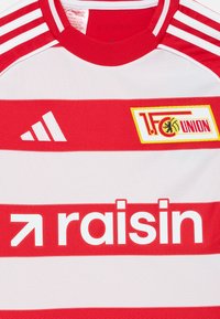 Rdeča in bela črtasta atletska majica s krožnim ovratnikom, ki vsebuje logotip "1.FC UNION" in belo grafiko "raisin", poleg tega pa tudi Adidasovo znamko.