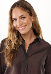 Camicia button-up marrone scuro con una texture liscia, dotata di colletto e vestibilità morbida. La camicia ha un leggero lucido e bottoni marroni.