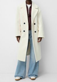 Crèmekleurige dubbele rij jas met grote zwarte knopen en zijzakken, gedragen over een bordeauxrode jas met rits, gecombineerd met wijd uitlopende spijkerbroeken.