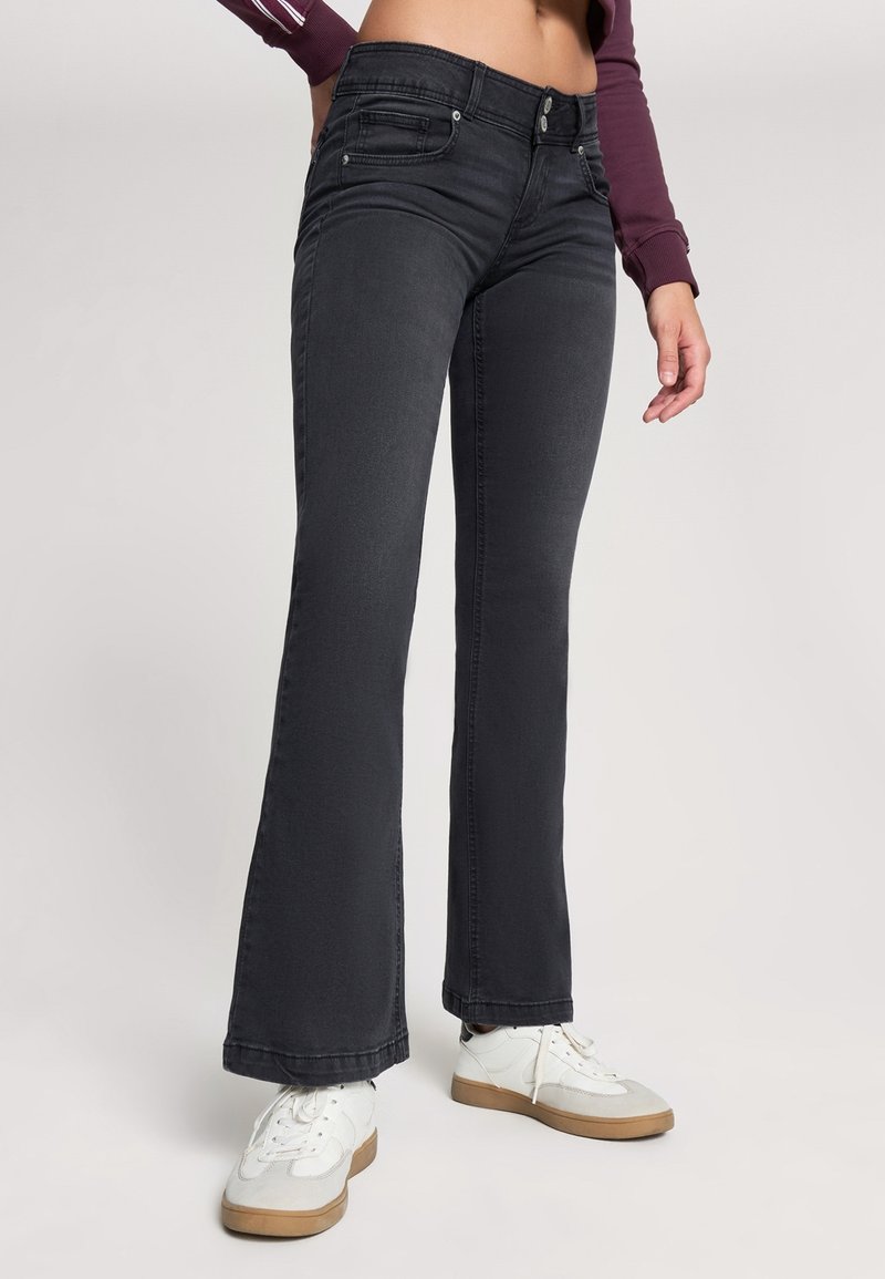 Czarne dżinsy w stylu dzwonów z średnim stanem, przednimi kieszeniami i subtelnymi detalami przeszycia, zestawione z białymi trampkami.