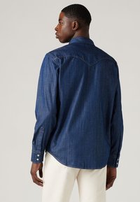Camisa de denim en azul oscuro con puños de botón y detalles en el yugo. Presenta un dobladillo recto y un pliegue en la espalda, mostrando una textura suave.