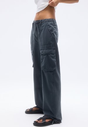 Persona che indossa pantaloni cargo larghi scuri e sandali neri, sollevando leggermente la maglietta bianca per mostrare l'addome.