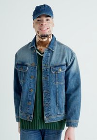 Denim jakna svijetloplave boje s dvije prednje džepove, uparena s tamnozelenim pletenim džemperom. Model nosi plavu kapu i traperice.