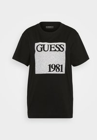 Svart bomulls t-shirt med ett silverfärgat grafiskt tryck med texten "GUESS 1981" i en rektangulär design. Standard rund hals och korta ärmar.