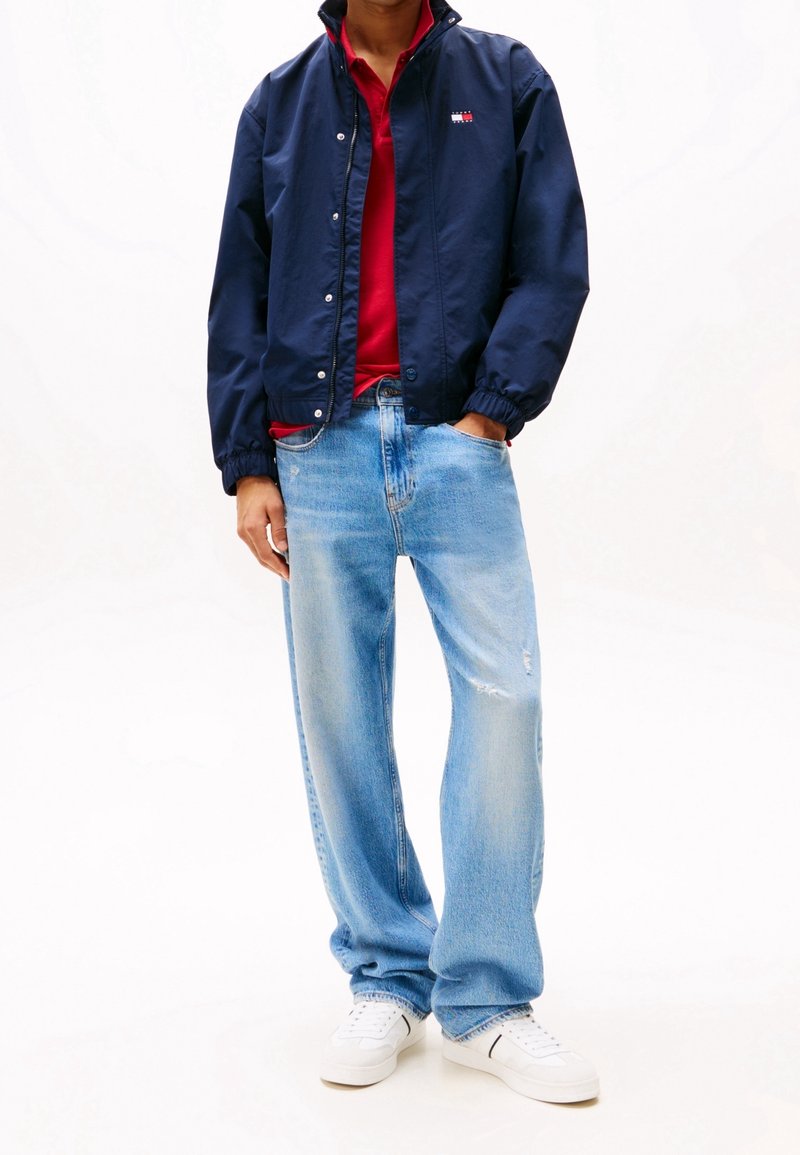 Personne portant une veste bleu marine par-dessus une chemise rouge, un jean large bleu clair et des baskets blanches, debout avec une main dans la poche.