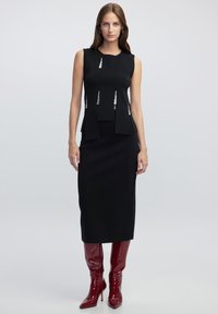 Zwarte mouwloze jurk met een aansluitende top, geplooide accenten en een rechte rok. Draag met glanzende bordeauxrode kniehoge laarzen.