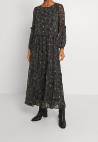 Lång, svart blommig klänning i lätt tyg med ett lager-på-lager design. Långa ärmar och volangdetaljer, med gröna och rödaktiga mönster.