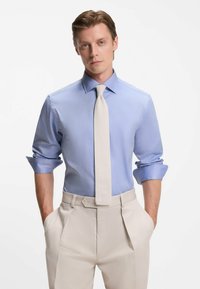 Camicia azzurro chiaro con maniche arrotolate, abbinata a una cravatta crema e pantaloni beige plissettati, con una texture di tessuto liscia.