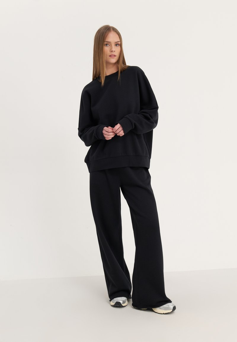 Ung kvinde står iført løs sort sweatshirt og brede sorte bukser med grå og hvide sneakers på en ensartet lys baggrund.