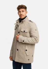 Manteau beige en mélange de laine avec un design croisé, grandes poches, boutons noirs et un col montant, présentant une silhouette structurée moderne.