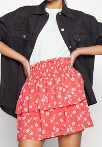Femme portant un t-shirt blanc, une veste noire ouverte à boutons, et une mini-jupe rouge à volants fleuris avec taille élastique, mains posées sur les hanches.