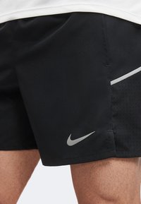 Shorts de sport noirs avec panneaux latéraux perforés et logo Nike réfléchissant, portés par une personne à la peau claire.