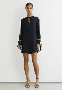 Tumši zila kleita ar garām piedurknēm un ar atslēgas cauruma formu pie kakla, rotāta ar baltām svītrām uz piedurknēm un saitēm aprocēs.