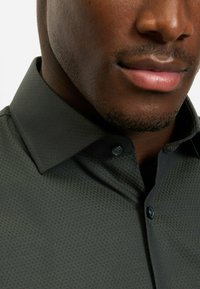 Dunkelgrünes, gemustertes Hemd mit strukturiertem Finish, ausgestattet mit einem klassischen Kragen und dunklen Knöpfen, aus der Nahaufnahme aufgenommen.