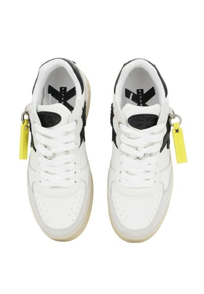 Een paar witte sneakers met zwarte accenten, witte veters, ecru zolen en gele labels bevestigd bij de enkels, gezien van bovenaf.