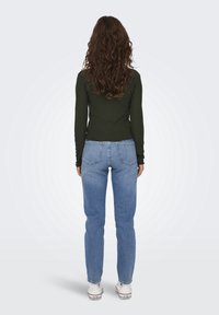 Frau mit langen lockigen Haaren, die ein dunkelgrünes Langarmshirt, blaue Jeans und weiße Turnschuhe trägt und mit dem Rücken zu uns auf einem schlichten Hintergrund steht.