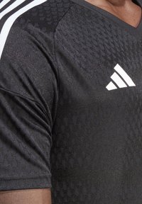 T-shirt de sport noir avec une surface texturée et un léger éclat. Il présente un col en V et des rayures blanches sur les épaules. Logo Adidas en blanc.