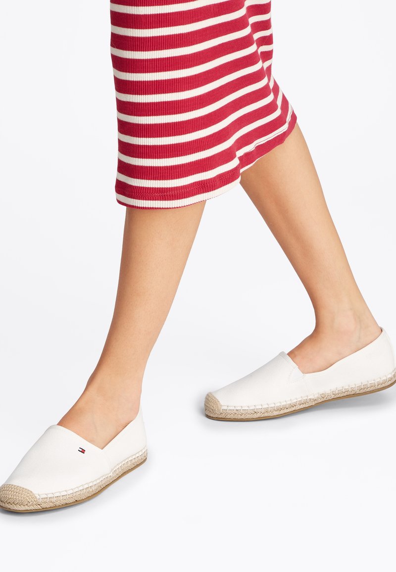 Espadrilles en toile blanche avec une semelle en jute tressé et un talon ouvert. Présentant un petit logo de marque sur le côté. Portées avec une jupe rayée rouge et blanche.