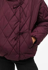 Kvalitetna maroon puf jakna s kapuljačom, koja ima vezanje na vezicu, bočne džepove i mekan, teksturiran materijal.