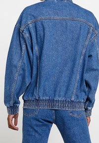 Person wearing a loose-fitting blue denim jacket and matching blue denim jeans, viewed from the back against a plain background.