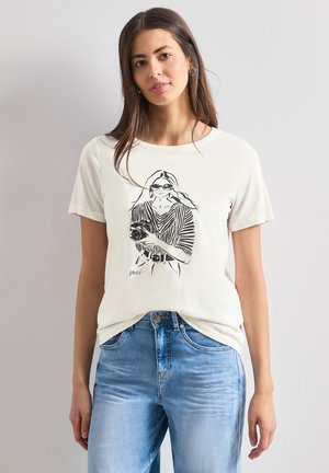 Frau mit langen braunen Haaren, die ein weißes T-Shirt mit einer schwarzen Skizze einer Frau hält, die eine Kamera hält, und blaue Jeans trägt, stehend vor einer schlichten Wand.