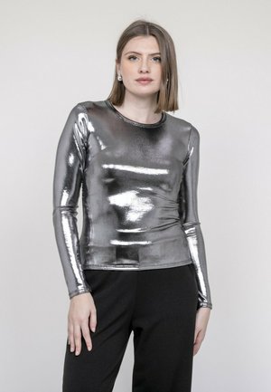 Femme aux cheveux mi-longs portant un haut métallisé argenté à manches longues et un pantalon noir, debout devant un fond clair uni.