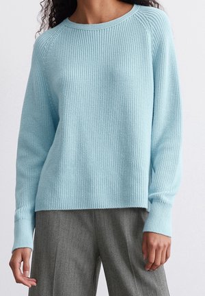 Kobieta ubrana w jasnoniebieski dzianinowy sweter z długimi rękawami i szare szerokie spodnie, stojąca na jednolitym białym tle.